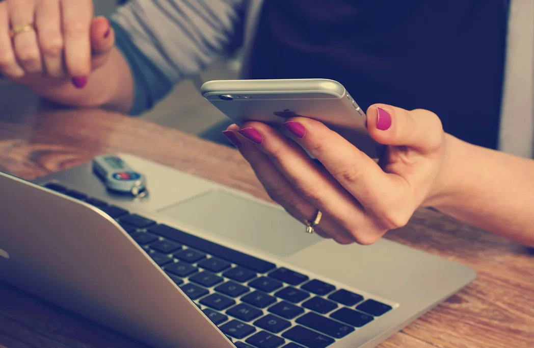Woman with a phone in her hand while sitting on a desk with a laptop